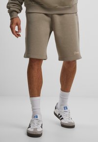 Beige bomullsshorts med avslappet passform, med ribbestrikket midjebånd, sidelommer og en liten logo detalj på kanten. Kombinert med hvite sokker og sneakers.