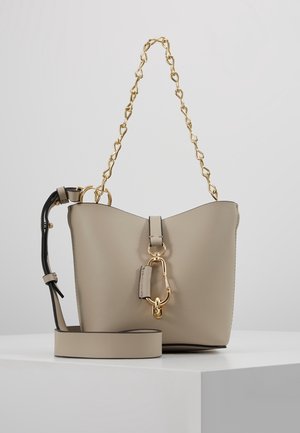 Sac seau beige avec bandoulière chaîne dorée, accent noir sur la poignée latérale et large fermoir doré à l'avant, présenté sur surface blanche.