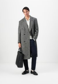Mann in einem grauen double-breasted Mantel, weißem Hemd, navy-blauen Hosen, schwarzen Slippern, der eine schwarze Leder-Umhängetasche hält und vor einer weißen Wand steht.