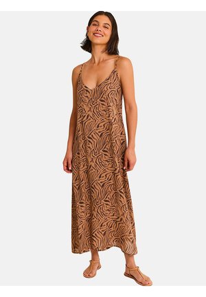 Femme portant une robe maxi longue sans manches à imprimé zèbre marron avec de fines bretelles et des sandales plates beige, souriante sur un fond uni.