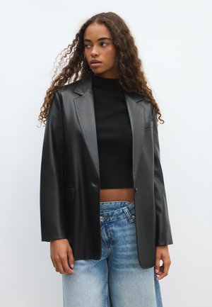 Femme aux cheveux bouclés portant un blazer en cuir noir par-dessus un crop top noir et un jean bleu clair, regardant sur le côté sur un fond uni.
