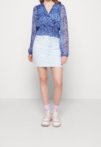 Blusa floreale blu con maniche lunghe trasparenti, abbinata a una mini gonna di jeans chiari con orli sfrangiati. Indossata con calzini rosa e sneakers chunky.