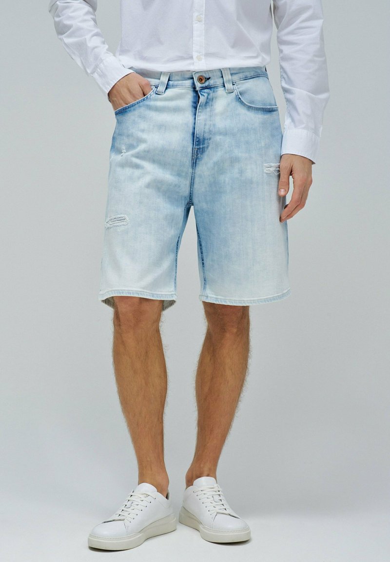 Pantalones cortos de mezclilla de lavado claro con un ajuste relajado, que presentan acentos desgastados y un dobladillo sin terminar, combinados con zapatillas blancas.