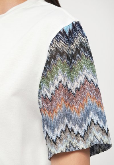 Λευκό βαμβακερό T-shirt με υφασμένη κοντομάνικη με πολύχρωμο ζιγκ-ζαγκ μοτί�βο, που περιλαμβάνει αποχρώσεις του μπλε, πράσινου, καφέ και πορτοκαλί.