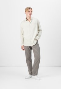 Lys beige skjorte med knappestolpe og en enkelt brystlomme, kombinert med grå kordfløyelsbukser og hvite sneakers, stående mot en ensfarget bakgrunn.