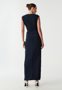 Granatowa, dopasowana sukienka maxi z krótkimi rękawkami, subtelnym połyskiem i zamkiem błyskawicznym z tyłu. Stylizowana z srebrnymi szpilkami i bransoletką.