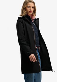 Abrigo largo negro con capucha, con cremallera frontal y puños elásticos. Se lleva sobre un suéter de punto de cable azul marino y unos vaqueros de corte claro.