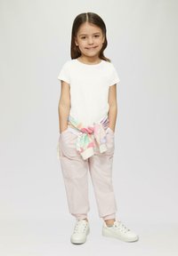 Weißes T-Shirt mit kurzen Ärmeln und kleinen farbigen Herzakzenten, pastellfarbenes gemustertes Sweatshirt um die Taille gebunden, hellrosa Hose, weiße Sneaker.