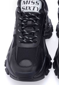 Sneaker sportive nere con tomaia in rete e pelle, punta rotonda, suola in gomma sollevata e lacci bianchi a contrasto con un motivo a scacchi.