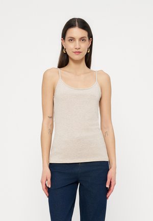 Femme aux longs cheveux noirs portant un haut bretelles fines beige, un pantalon bleu et des boucles d'oreilles créoles dorées, debout devant un fond blanc uni.