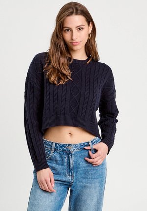 Marineblauwe cropped kabelgebreide trui met ribbelhals en -manchetten, voorzien van een textuur van diamanten en kabelpatronen, gecombineerd met lichtblauwe jeans.