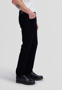 Personne portant un pantalon noir et des bottes en cuir noir, avec la main dans la poche et un tatouage visible sur l'avant-bras sur un fond uni.