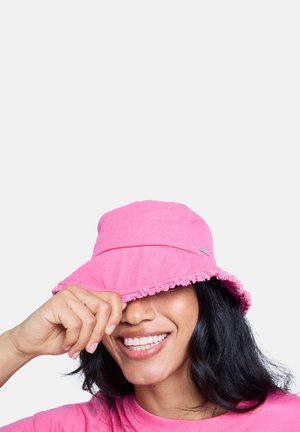 Sombrero tipo bucket rosa con ala deshilachada, hecho de tela. Un pequeño logo es visible en el lateral. La persona que lo lleva sonríe mientras se cubre parcialmente el rostro.