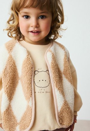Chaleco de forro polar beige con patrón de diamantes sobre una sudadera crema que presenta un oso de dibujos animados y el texto "¡HOLA! ES UN NUEVO DÍA." Pelo rizado.