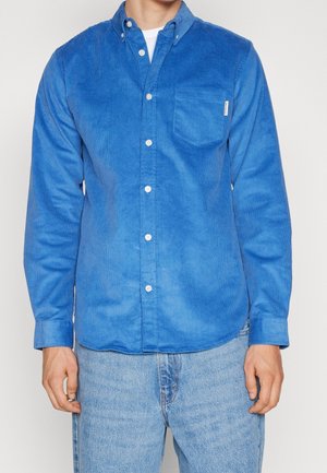 Hombre vestido con camisa de pana azul brillante de botones sobre camiseta blanca y jeans de mezclilla azul claro, de pie contra un fondo liso.