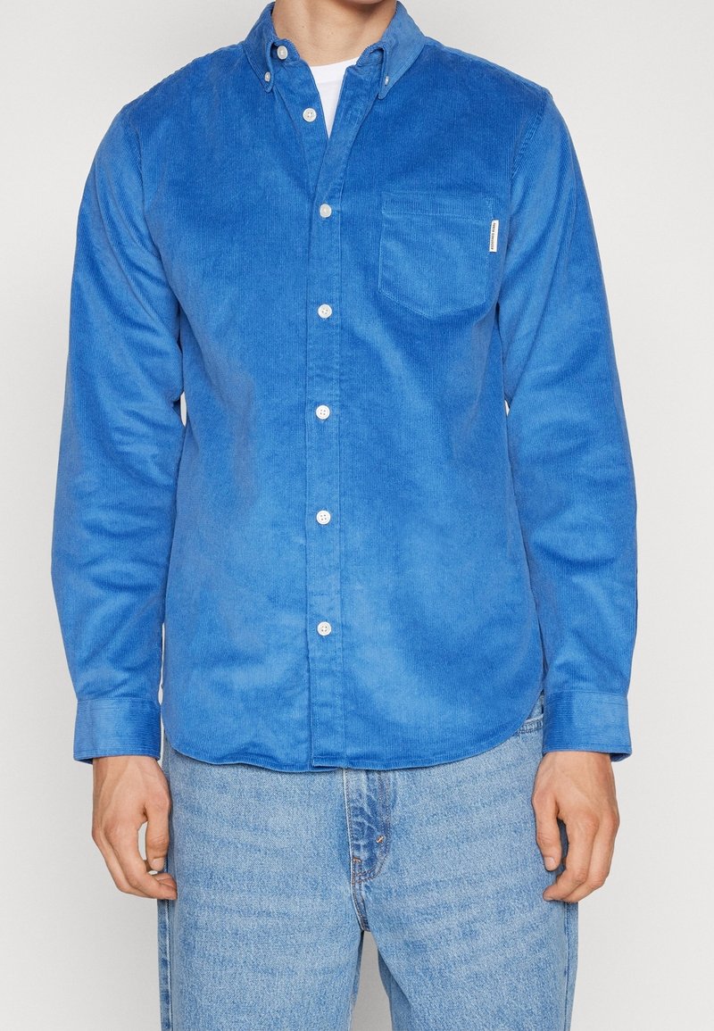 Homme portant une chemise en velours côtelé bleu vif boutonnée sur un t-shirt blanc et un jean en denim bleu clair, debout devant un fond uni.