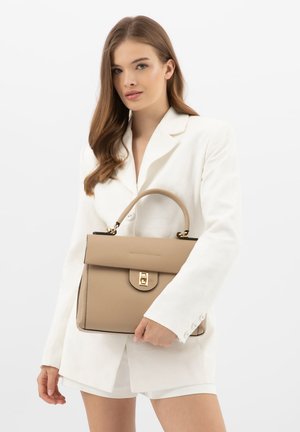 Žena v bílém saku a šortkách drží béžovou koženou kabelku s horním úchopem a zlatou sponou.
