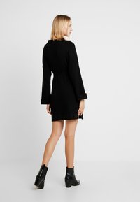 Zwarte gebreide jurk met lange mouwen en een ceintuur om de taille. Heeft een geribbelde textuur en een lengte boven de knie, gecombineerd met glanzende enkellaarsjes.