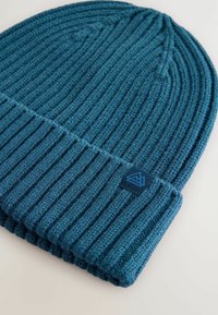 Cappello in maglia a coste di colore verde scuro. Presenta un orlo ripiegato e una piccola etichetta blu con un design di montagna sul lato.