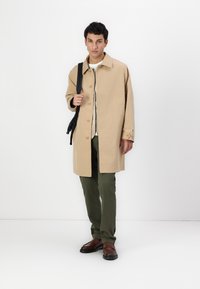 Cernă de trench bej cu nasturi mari, buzunare laterale și o croială lejeră. Asortat cu pantaloni oliv și pantofi brown slip-on.