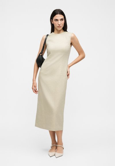 Γυναίκα που φοράει μπεζ μίντι φόρεμα χωρίς μανίκια, κρατάει μαύρη τσάντα ώμου, στέκεται με το ένα χέρι στη μέση και φοράει ανοιχτόχρωμα μυτερά παπούτσια.