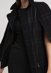 Manteau en laine à carreaux noirs et foncés avec fermeture éclair, poches zippées et col montant. Comprend des capes aux épaules et une finition lisse et texturée.