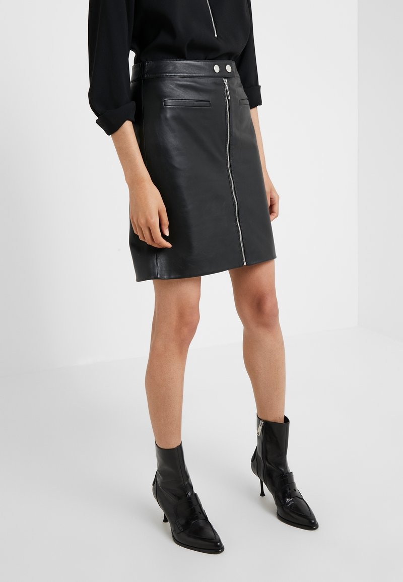 Femme portant une mini-jupe en cuir noire avec une fermeture éclair et des boutons à l'avant, associée à des bottines noires à talons hauts et un haut noir à manches longues.
