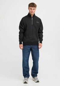Sudadera negra con cremallera en el cuello, fabricada en una mezcla de algodón. Presenta puños y dobladillo de canalé, con un pequeño logotipo "A.R.C." en el pecho. Llevada con jeans azules.