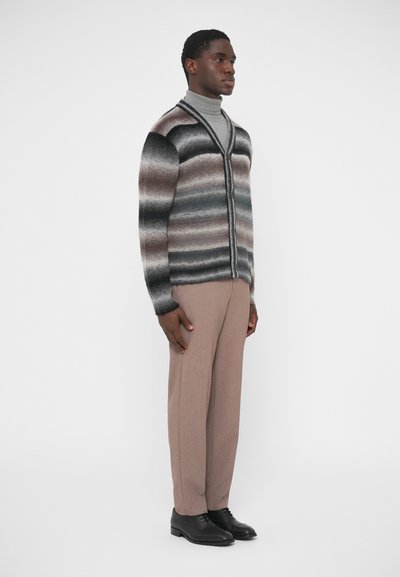 Reljeefne kardigan, millel on halli, pruuni ja musta triibulised üleminekud, V-kaeluse ja nööbitava eesosaga. Sobitatud kohandatud beežide pükstega ja mustade kingadega.