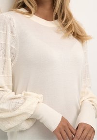 Žena na sobě má bílý pletený svetr s detailními krajkovými balónovými rukávy a žebrovanými manžetami, ruce jemně sevřené vpředu.