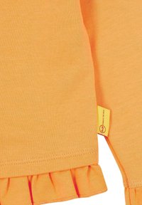 Oranje katoenen shirt met een gerimpelde onderrand, een soepele textuur en een gele label aan de zijkant met rode tekst, die merkinformatie aangeeft.