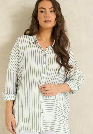 Vrouw met lang golvend haar, gekleed in een loszittend wit overhemd met dunne verticale groene strepen, staand tegen een effen beige achtergrond.