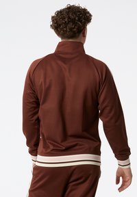 Hnědá sportovní bunda s vysokým límcem a pruhovanými manžetami. Má hladkou texturu a žebernaté akcenty v bílé a růžové barvě.