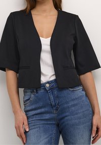Svart kort jacka med breda ärmar, öppen front och två små fickor, i kombination med en vit topp och blå denimjeans.