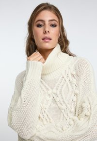 Flauschiger weißer Strickpullover mit hohem Rollkragen, ausgestattet mit Zopfmustern und strukturierten Bommel. Dicke gerippte Bündchen und Saum.