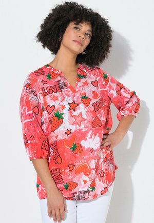 Frau mit lockigem Haar, die eine leuchtend pinke Bluse trägt, verziert mit Erdbeeren, Herzen, Sternen und dem Wort "LOVE", kombiniert mit weißen Hosen.