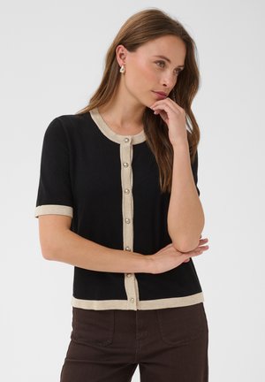 Femme aux longs cheveux bruns porte un cardigan noir à manches courtes avec finition beige et boutons, un pantalon marron et des boucles d’oreilles en perles, touchant son menton.