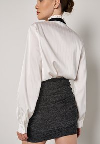 Blusa blanca a rayas con puños fruncidos, combinada con una minifalda negra ceñida y brillante que presenta detalles de fruncido en los lados.
