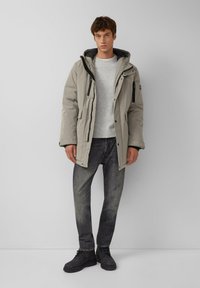 Grauer Parka mit Kapuze, ausgestattet mit Reißverschlusstaschen und einem strukturierten Pullover darunter. Schwarze Jeans und dunkle Stiefel vervollständigen das Outfit.