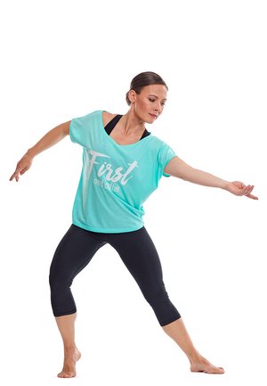 Light turquoise short-sleeve top with white text, paired with black cropped leggings; model in dynamic pose, barefoot.