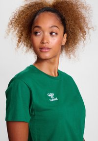 Camiseta de algodón verde con cuello redondo y mangas cortas, que presenta un logo blanco impreso en el área del pecho izquierdo. Textura suave.