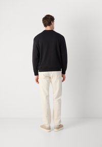 Sweatshirt noir avec des poignets et un ourlet côtelés, associé à un pantalon beige clair. Modèle de dos sur un fond blanc uni.