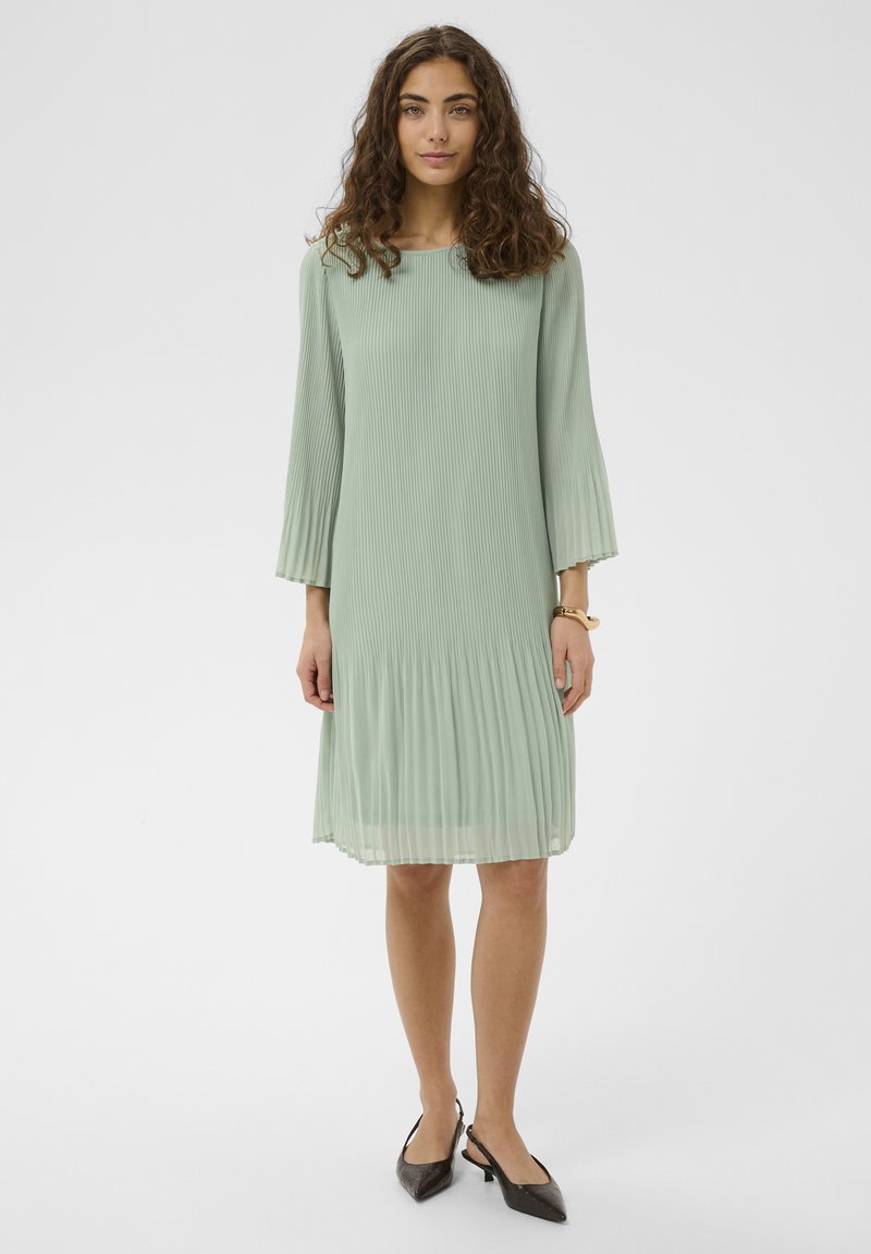 Ženska s kodrastimi lasmi, oblečena v svetlo zeleno plisirano obleko dolžine do kolen in temne ukazane črpalke s trakom za peto, stoji na belem ozadju.