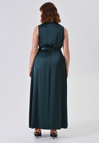 Donna con capelli castani lunghi fino alle spalle, che indossa un abito maxi senza maniche di raso verde scuro e scarpe con tacco nere, girata di spalle.