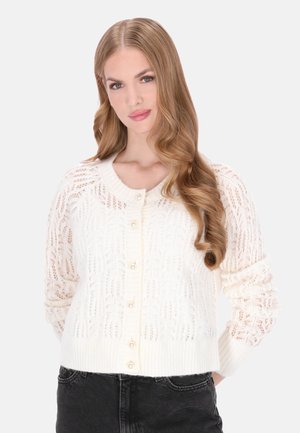 Gestrickter weißer Cardigan mit einem Spitzenmuster, gerippten Bündchen und Saum sowie Perlenknöpfen vorne. Über einem schwarzen Outfit getragen.