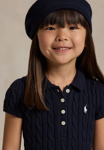 Lachend meisje met steil donker haar, met een marineblauwe gebreide muts en een poloshirt met korte mouwen en kabelpatroon, voorzien van witte knopen en logo.