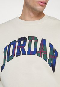 Gros plan d'une personne portant une chemise beige avec « JORDAN » en grandes lettres à motif à carreaux sur la poitrine.