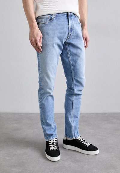 Jasnoniebieskie dżinsy o slim fit, z delikatnym wyblaknięciem, pięcioma kieszeniami i kontrastowym stębnowaniem, w połączeniu z czarnymi sneakersami.