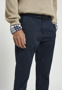 Chinos navy en coton, avec une texture lisse et une coupe classique. Les poignets révèlent un motif à carreaux bleu et blanc.