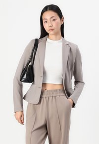 Beige sako s kontrastními klopami přes bílý žebrovaný top, v kombinaci s béžovými kalhotami s elastickým pasem. Černá kabelka se zlatým zipem.